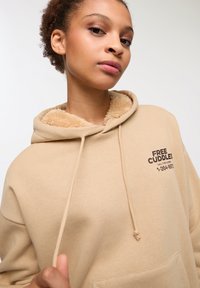 Brązowa bluza z kapturem z pluszowym, beżowym wnętrzem, widocznymi sznurkami do ściągania i przednią kieszenią. Nadrukowany tekst na czarno: "DARMOWE PRZYTULANIE, Zadzwoń pod ten numer."