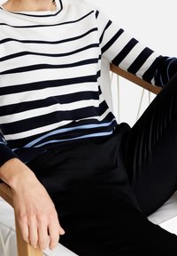 Navy- und weiß gestreifter Pullover mit einem hellblauen Akzent am Saum, aus weichem Stoff. Kombiniert mit schwarzen Hosen. Lässiger Sitzbereich im Hintergrund.