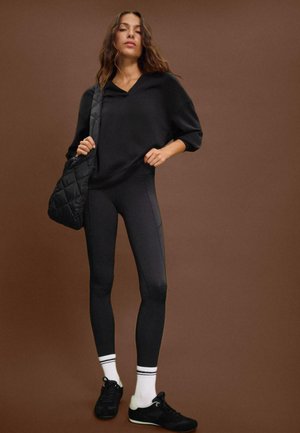 Femme se tenant contre un fond marron, portant un hoodie noir, des leggings noirs, des chaussettes blanches avec des rayures noires, des baskets noires, et portant un sac matelassé noir.