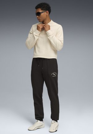 Beige lange mouw top met verticale strepen, gecombineerd met zwarte joggingbroek met logo-detail, en witte sneakers. Model draagt een zonnebril.