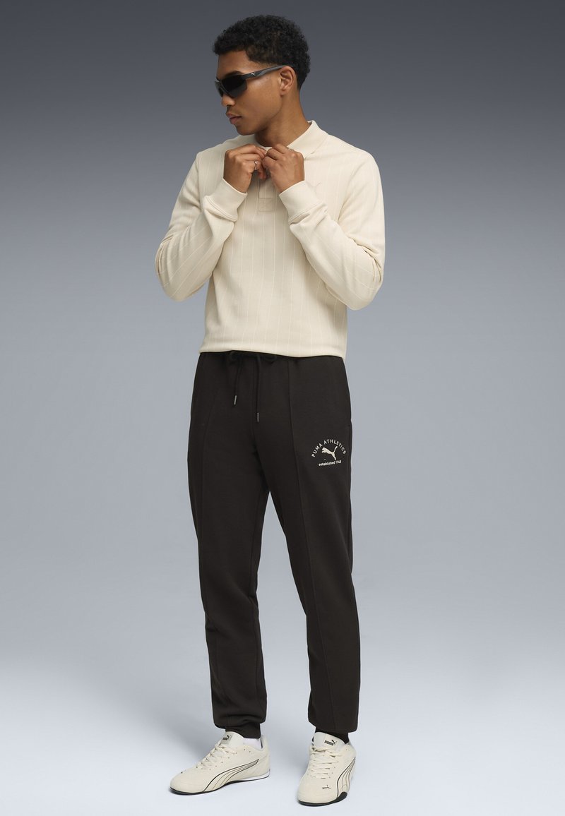 Beige langærmet top med lodrette striber, parret med sorte sweatpants med logodetalje, og hvide sneakers. Modellen bærer solbriller.