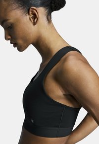 Soutien-gorge de sport noir avec un design ajusté, des bretelles larges et une texture lisse. Affiche un branding minimal avec une étiquette sur le devant.