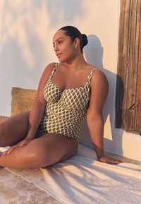 Costume intero con motivo a diamanti verdi su sfondo crema, con spalline larghe e tessuto strutturato, indossato da una figura seduta.