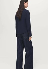 Navy blouse with long sleeves, featuring a rounded hem and gold button accents. Paired with high-waisted dark denim, wide-leg jeans.