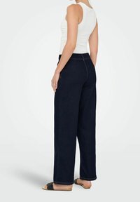 Kraftiga mörk denimjeans med hög midja, med kontrasterande sömmar och bakfickor. Matcher med en vit linne och sandaler.