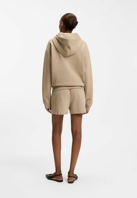 Frau mit zu einem Dutt gebundenem Haar, die einen beigen Kapuzenpullover und passende Shorts trägt, steht mit dem Rücken zum Betrachter vor weißem Hintergrund.