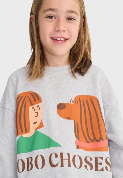 Lächendes junges Mädchen, das ein graues Sweatshirt mit einer Illustration eines Mädchens und eines Hundes trägt, die sich gegenüberstehen, und dem Text "BOBO CHOSES".