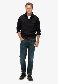 Zwarte button-up shirt met twee borstzakken, gecombineerd met blauwe denim jeans. Model draagt een bruine riem en zwarte veterschoenen.