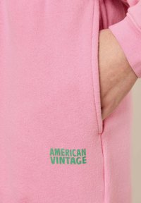 Sweat à capuche rose avec une texture douce, arborant un logo vert "AMERICAN VINTAGE" en bas à gauche et une main insérée dans la poche.