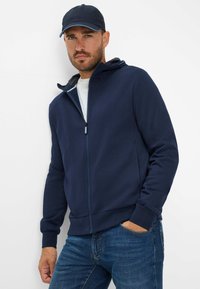 Felpa con zip di colore blu navy realizzata in tessuto morbido, dotata di cappuccio con cordino, tasche frontali e polsini a coste, abbinata a jeans di denim blu.