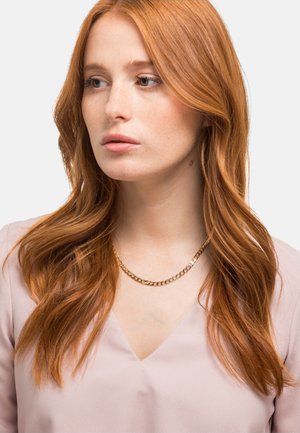 Collier en chaîne dorée à deux couches, porté sur un chemisier rose clair à col en V. Texture lisse, style minimaliste.