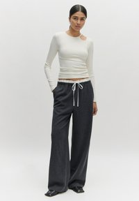 Langsleeve ivoorkleurige top met een zijuitsnijding, gecombineerd met losvallende donkergrijze broek met een elastische tailleband en witte trekkoord.