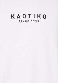 T-shirt bianco con stampa di testo nera che dice "KAOTIKO" seguito da "SINCE 1999". Materiale in cotone con una texture liscia.