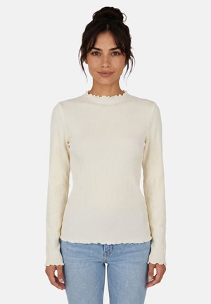 Donna con i capelli scuri raccolti indossa una maglia crema aderente a maniche lunghe con bordi arricciati e jeans azzurri chiari, in piedi contro uno sfondo bianco.