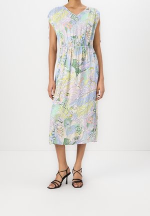 Femme portant une robe midi de couleur pastel avec des motifs floraux abstraits et du texte, associée à des sandales noires à talons hauts à brides.
