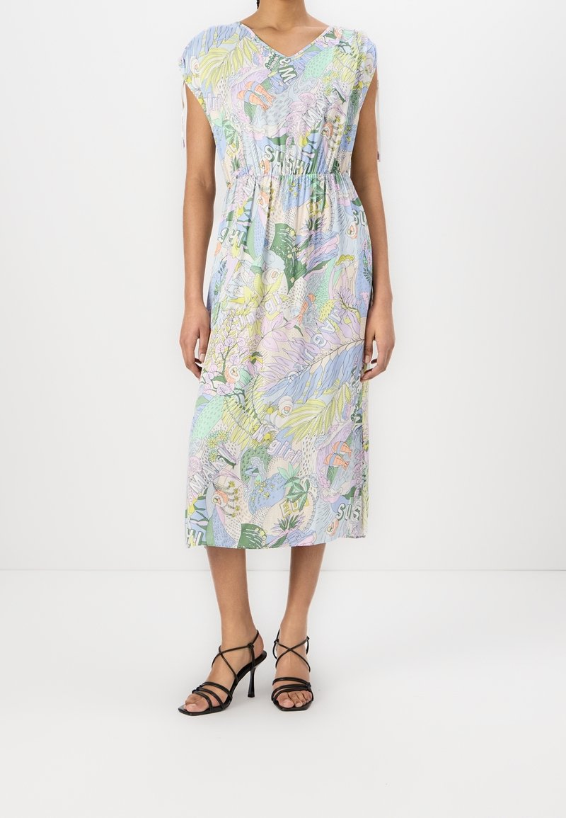 Femme portant une robe midi de couleur pastel avec des motifs floraux abstraits et du texte, associée à des sandales noires à talons hauts à brides.