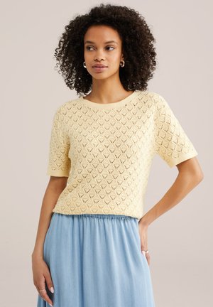 Donna con capelli ricci che indossa una maglia a maniche corte di colore giallo chiaro e una gonna a vita alta azzurro chiaro, in piedi con una mano sul fianco.