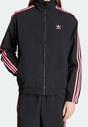 Personne portant un survêtement noir Adidas avec des bandes roses sur les manches et le pantalon, veste zippée, main dans la poche.