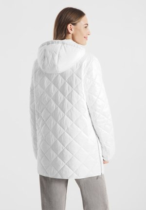 Vrouw die een witte gewatteerde jas met capuchon draagt, vanuit de achterkant gezien, met lichtgrijze broek tegen een effen achtergrond.