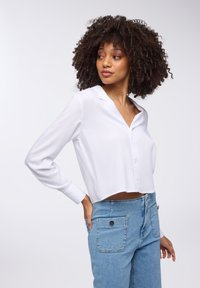 Camicetta bianca corta con bottoni sul davanti e colletto, abbinata a jeans a vita alta azzurri chiari con tasche posteriori e dettagli in metallo argentato.