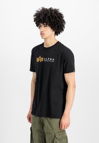 Μαύρο βαμβακερό t-shirt με λευκό και χρυσό λογότυπο "Alpha Industries". Κλασικός σχεδιασμός με στρογγυλή λαιμόκοψη, κοντά μανίκια και άνετη εφαρμογή.