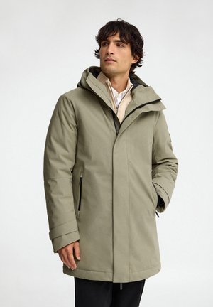 Uomo con parka kaki con cappuccio sopra un maglione beige e camicia bianca, in piedi con una mano in tasca, che guarda di lato.