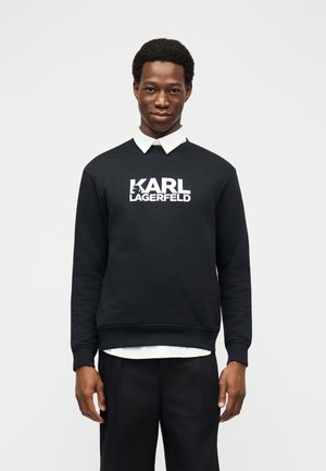 Young man wearing a black sweatshirt with white "KARL LAGERFELD" text and a white collared shirt underneath, standing against a plain background.