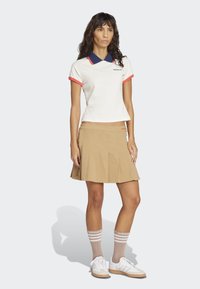 Biely polo tričko s námorníckymi a koralovými akcentmi, béžová plisovaná sukňa a kožené hnedé ponožky do kolien s bielymi prúžkami. Biely športový obuv dokončuje outfit.