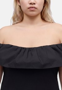 Femme à la peau claire et aux cheveux blonds portant un haut noir épaules dénudées, montrant son cou, ses épaules et le haut de sa poitrine.