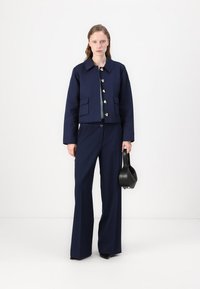 Marineblauwe cropped jacket met ronde kraag, vier zakken en gouden knopen, gecombineerd met wijde marineblauwe broeken en een zwarte handtas.