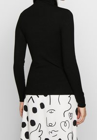 Kvinna som bär en svart långärmad polotröja och en vit kjol med abstrakt svart linjekonst och prickmönster, sedd bakifrån.