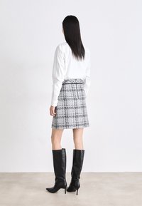 Jupe écossaise grise avec une forme ajustée et des plis, associée à des bottes en cuir noires à talons haut qui montent jusqu'aux genoux et une chemise blanche à col.