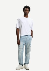 Bjela kratka majica s kratkim rukavima, opuštenog kroja; svijetloplave cargo traperice s bočnim džepovima; bijele tenisice; strukturirana kosa. Jednostavna pozadina.