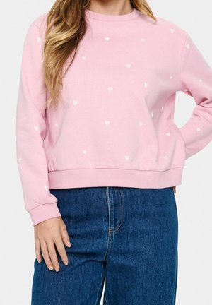 Nainen, joka pukeutuu vaaleanpunaiseen collegepaitaan, jossa on pieniä valkoisia sydämiä, ja korkeavyötäröisiin sinisiin farkkuihin yksinkertaisen taustan edessä.