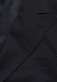 Tkanina czarnej marynarki, charakteryzująca się gładką teksturą, ostrymi klapami i małą kieszonką. Widoczne szwy podkreślają dopasowaną konstrukcję.