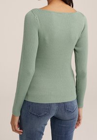 Kvinna iklädd en figurnära ribbad grön långärmad topp i salvia, som är ihop med blå jeans, sedd bakifrån mot en neutral bakgrund.