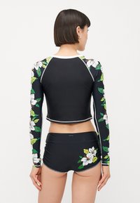 Black rash guard with long sleeves and floral print in white and green. Paired with high-waisted black shorts featuring matching floral accents.