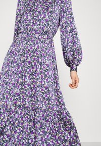 Maxi šaty s květinovým potiskem v purpurové, fialové a žluté na tmavém pozadí, s dlouhými nafouknutými rukávy a přestřiženým pasem.