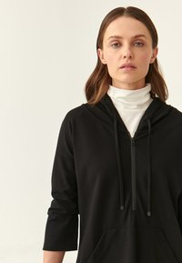 Hoodie noir en tissu doux, avec un devant zippé, des cordons de serrage ajustables, une poche avant et des manches trois-quarts.