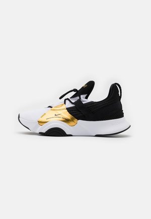 Baskets slip-on blanches, noires et dorées avec empeigne en mesh, bandes élastiques et languette au talon, avec le texte « Groove Sweat Repeat. »