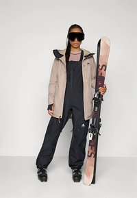 Γυναίκα που φορά γυαλιά σκι, μπεζ μπουφάν, μαύρη στολή σκι και μπότες, κρατώντας ένα ροζ και μαύρο σκι κατακόρυφα δίπλα της.