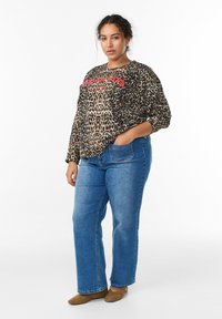 Žena v leopardom vzorovanom tričku s dlhým rukávom s červenými písmenami, v modrých džínsoch a hnedých topánkach, stojaca s jednou rukou vo vrecku na bielom pozadí.