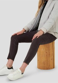 Osoba oblečená v černých kalhotách, bílém saku a bílých teniskách sedí na dřevěném válcovém stoličce na jednobarevném pozadí.