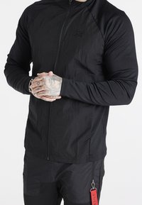 Svart zip-up jacka med texturerad tyg, raglanärmar och broderad logotyp. Funktioner inkluderar en röd etikett bifogad till byxor med dragkedja.