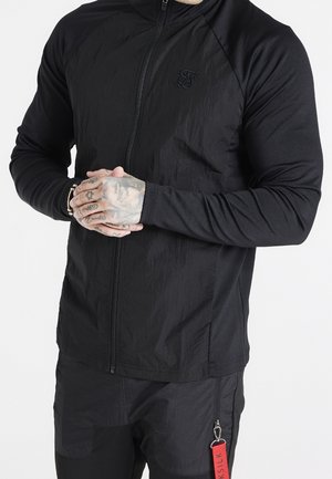 Chaqueta negra con cremallera, fabricada con un tejido texturizado, mangas raglán y un logotipo bordado. Presenta una etiqueta roja unida a los pantalones con una cremallera.