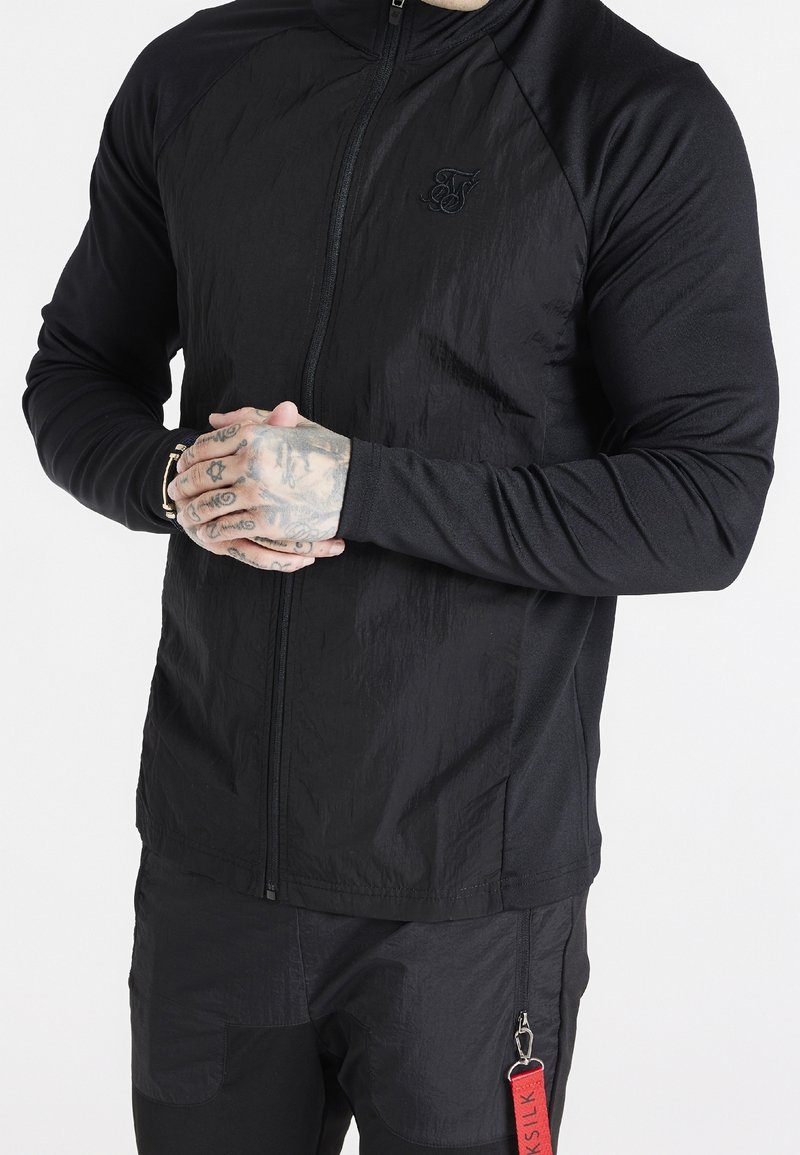 Svart zip-up jacka med texturerad tyg, raglanärmar och broderad logotyp. Funktioner inkluderar en röd etikett bifogad till byxor med dragkedja.