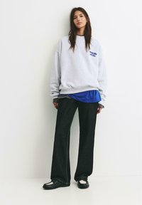 Felpa grigia con testo blu, indossata sopra una camicia blu, abbinata a jeans larghi scuri e mocassini neri. Design semplice e vestibilità rilassata.