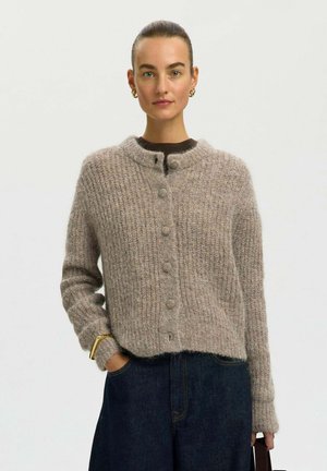 Femme aux cheveux tirés en arrière, portant un pull gris douillet boutonné, un jean foncé, des boucles d'oreilles en or et un bracelet en or, tenant un sac à main.