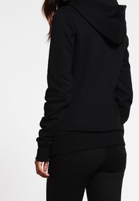 Osoba v přiléhavé černé mikině s kapucí a černých kalhotách, zobrazená zepředu na jednobarevném bílém pozadí.