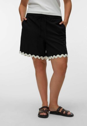 Personne portant un short noir à cordon avec un ourlet blanc festonné et des sandales noires plates, les mains dans les poches, debout devant un fond uni.